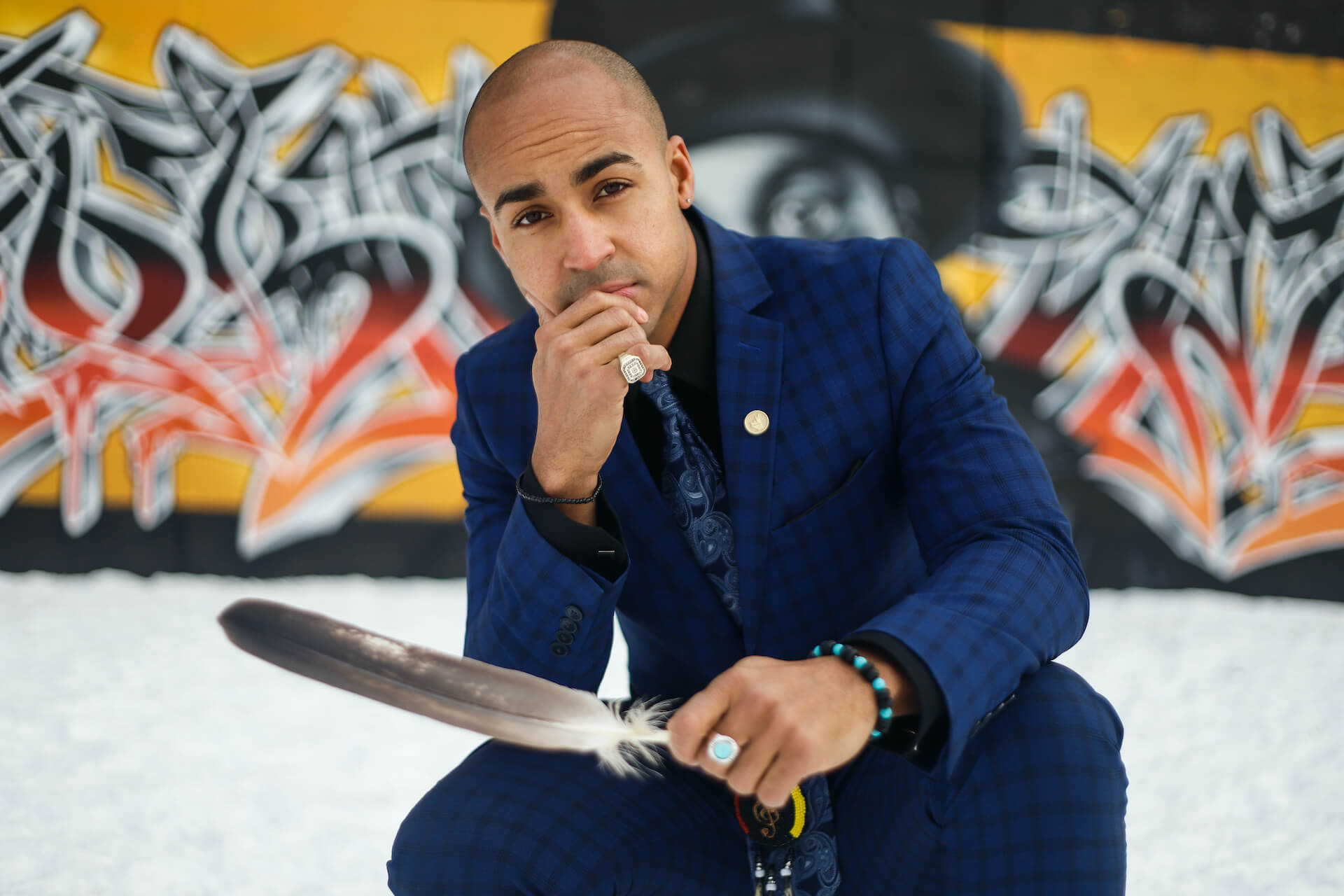 Close-up photo of Justin in a suit, in front of a wall of graffiti, holding a feather